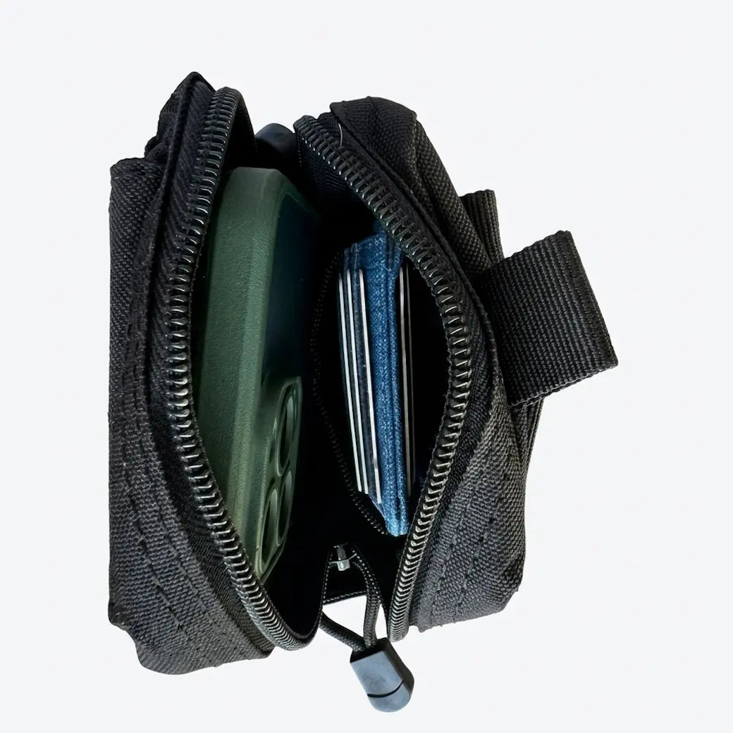 A black nylon everyday carry holder pouch shown open, displaying its interior compartments with a green phone case, cards, and a black strap against a light gray background.