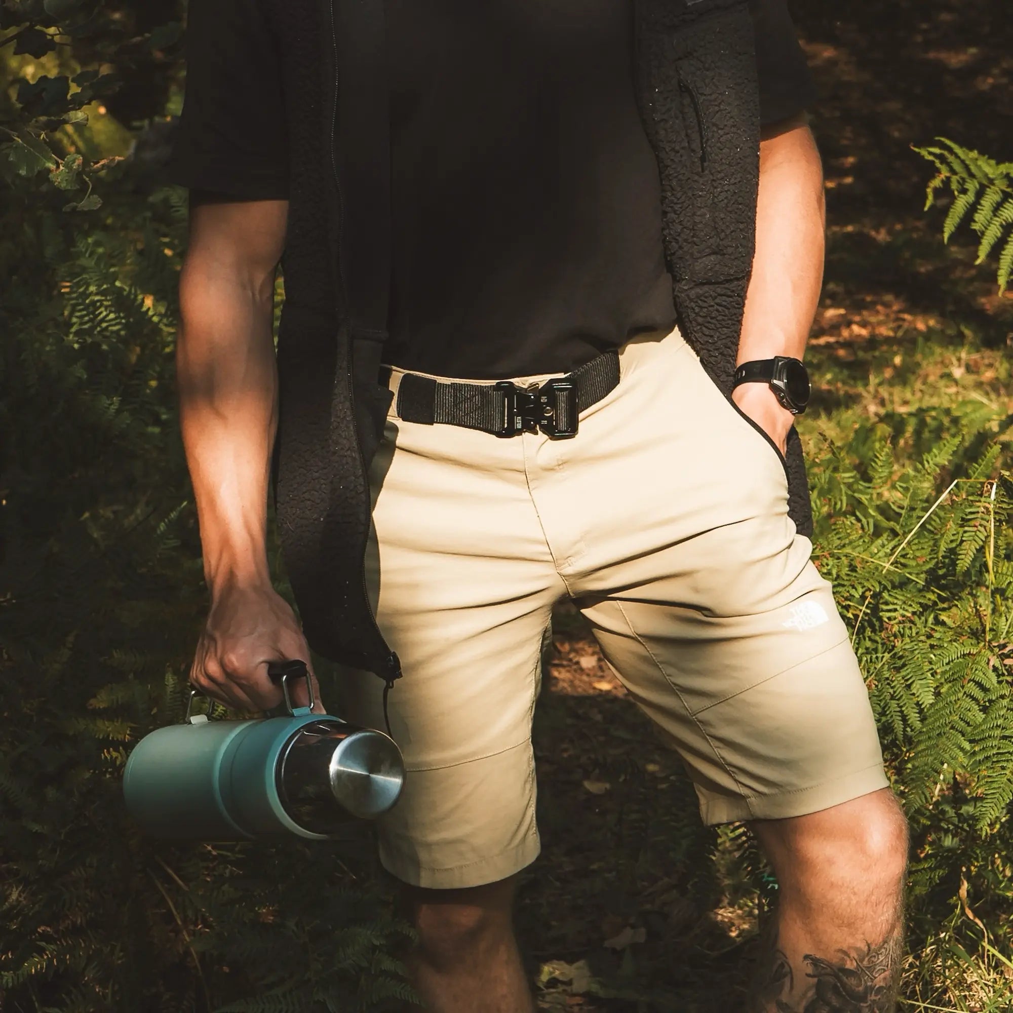 Man amongst ferns wearing a black Holsta Belt, beige shorts and black t-shirt and fleece while holding a blue flask