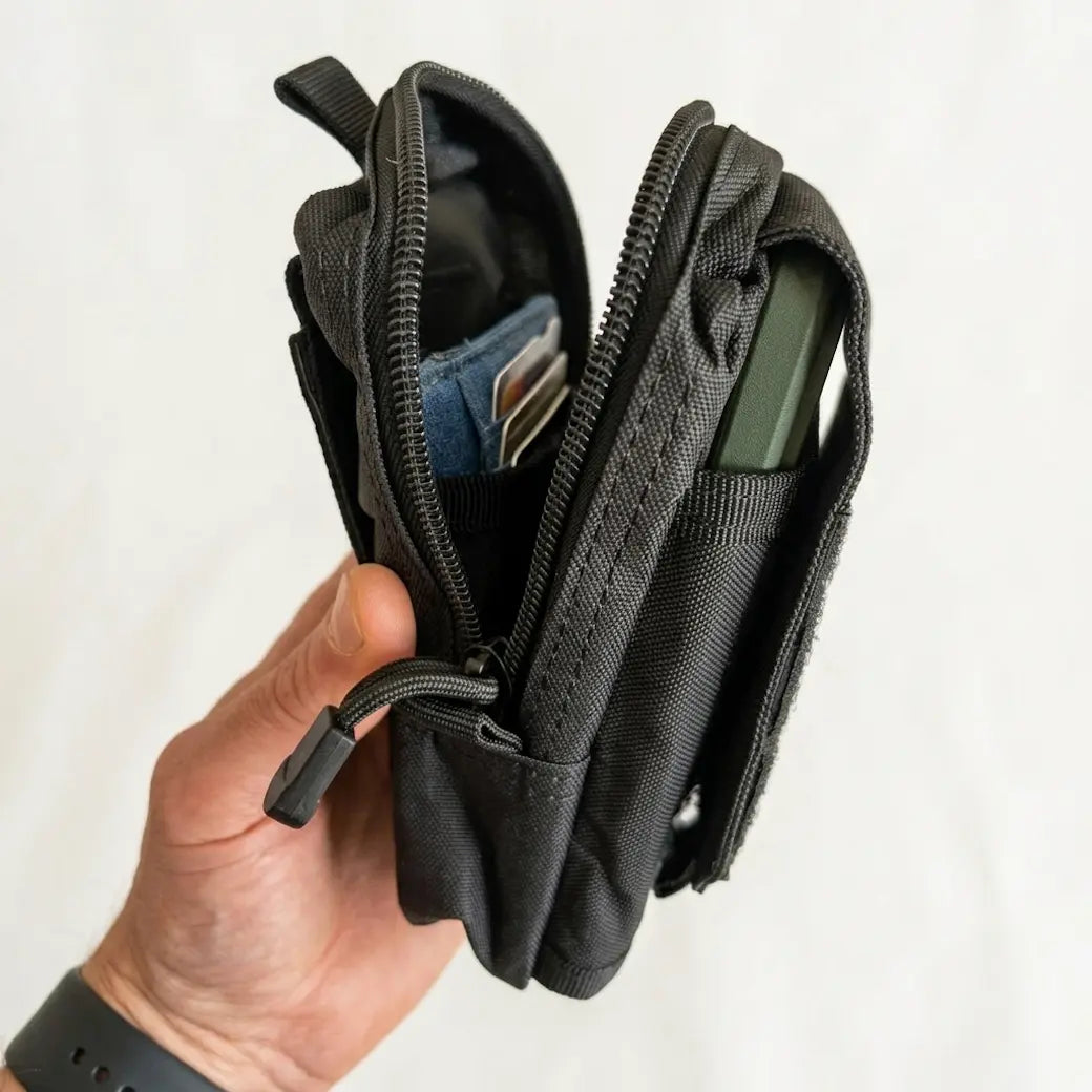 A black nylon utility pouch or small wallet being held open by a hand, showing interior compartments, zipper details, and card slots against a plain light background.