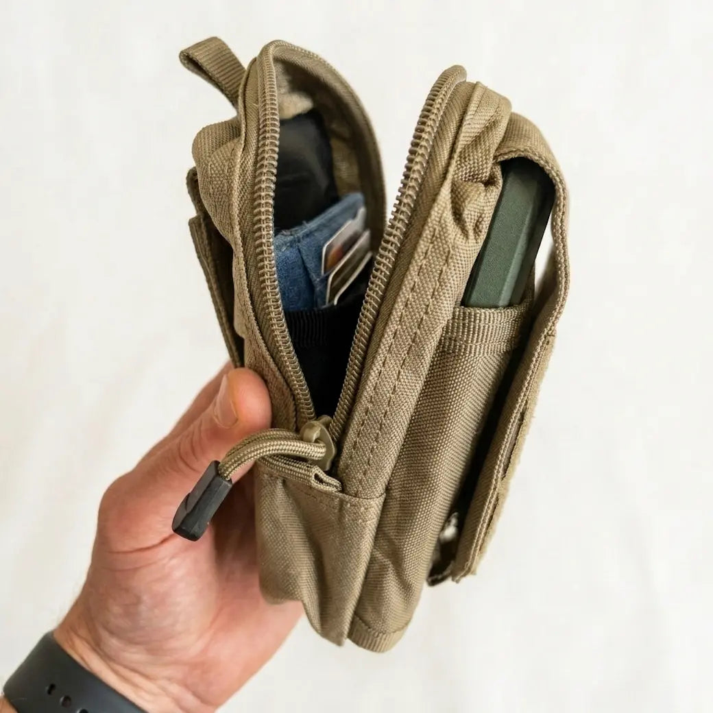 A hand holding a small tan canvas zip pouch wallet with an open zipper, showing card slots and a phone pocket, against a plain white background.