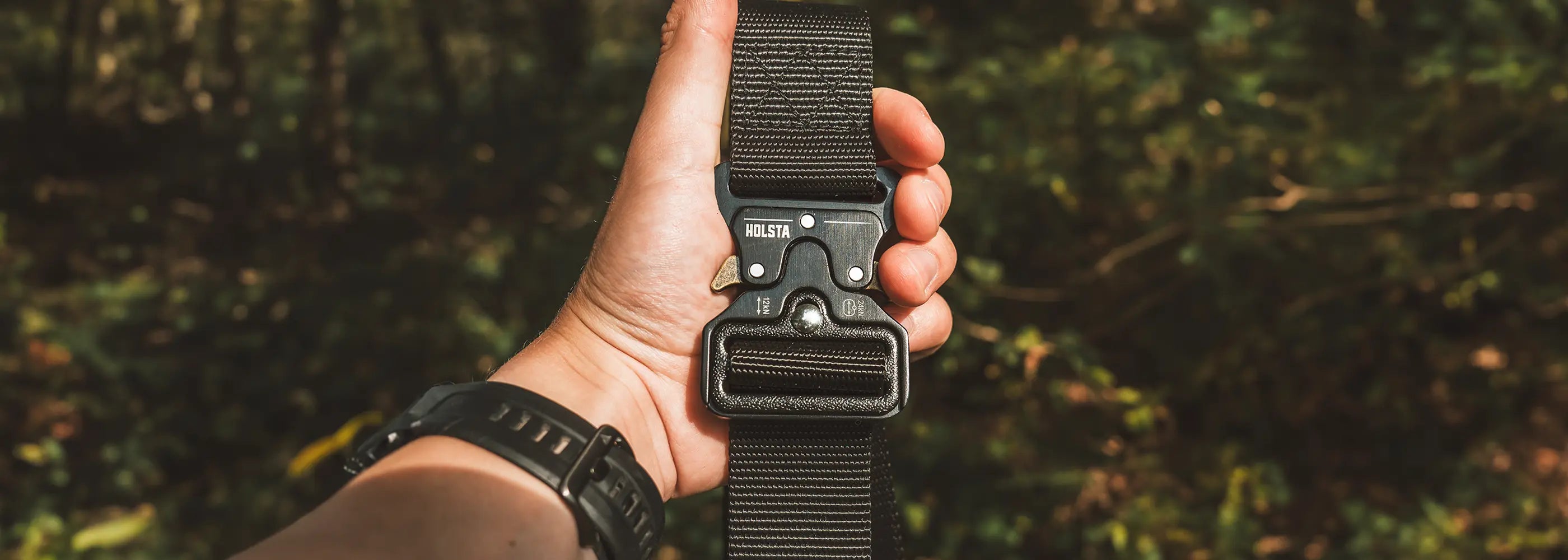 Hand holding up black Holsta Belt outdoors with blurry plants behind