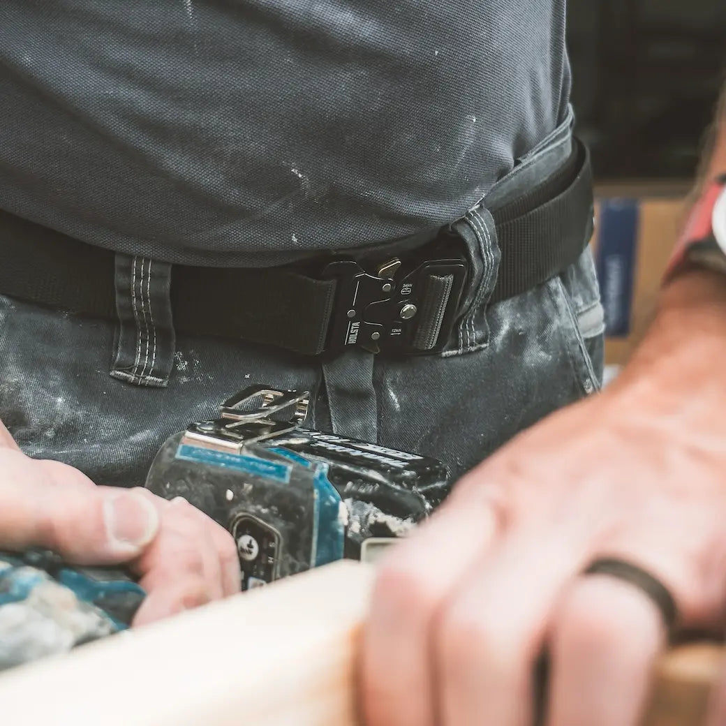 A close-up of a person wearing a black tactical belt with a black buckle, holding a blue and black cordless tool in a workshop setting.
